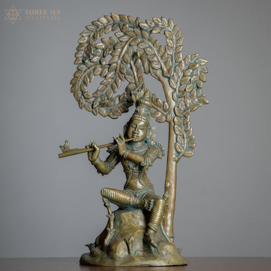 Bronze sculpture of Krishna playing a flute under a tree on a gray background